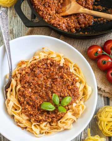 A white bowl of tagliatelle pasta topped with meat-like crumbles in bolognese sauce. Leftover bolognese in the cast iron skillet is right next to it.