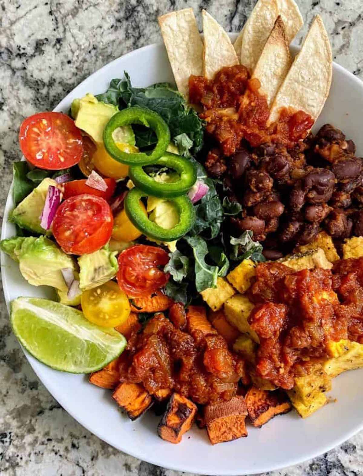 A white bowl with orange tofu cubes, red sauce, beans in sauce, chili pepper slices, avocado, cherry tomatoes and lime.