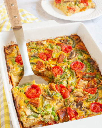 Cherry tomatoes, mushroom slices, bell pepper slices on top of a yellow cake-like dough is in a white casserole dish. A slice served on a white plate is in the background