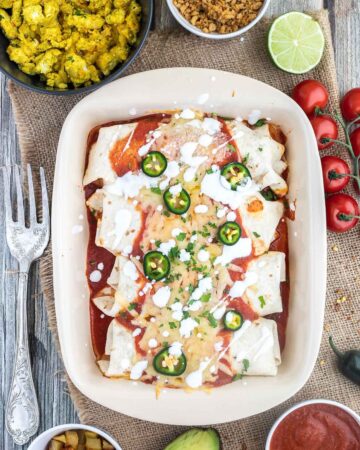 4 tortilla wraps topped with red sauce, melted cheese, sour cream, green chili peppers and fresh chopped herbs tucked closely together in a ceramic pan. Leftover filling ingredients are next to the pan in small bowls.