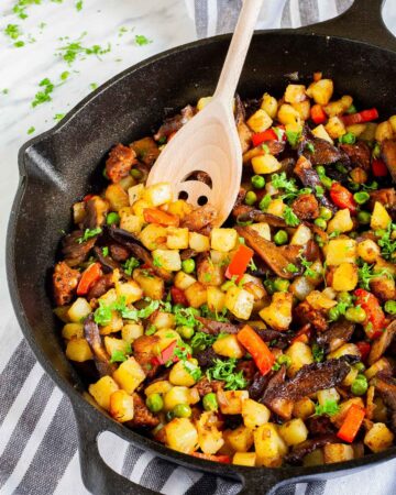 Black cast iron skillet from above with a wooden spatula and colorful chopped veggies like green peas, red bell pepper, yellow potatoes, brown mushroom, fresh parsley