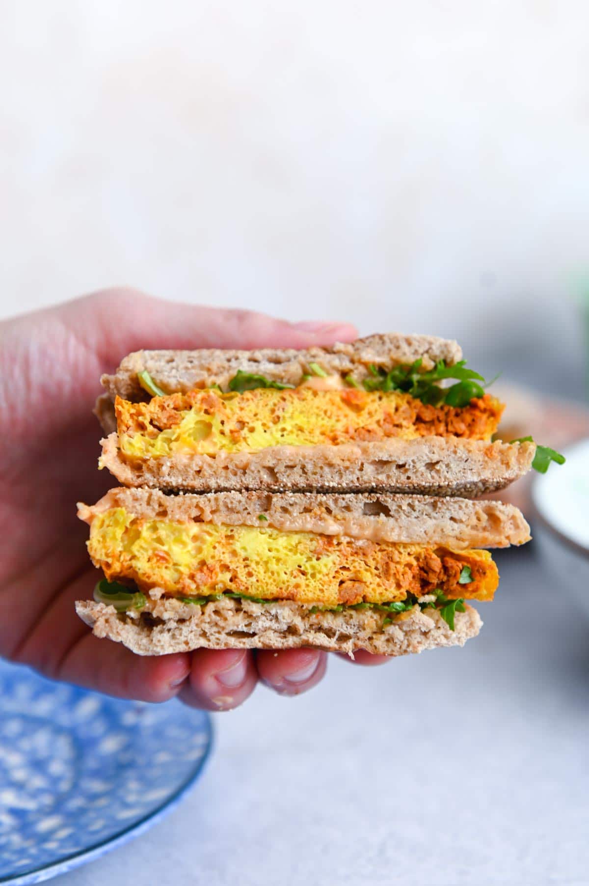 A hand is holding two small sandwiches with an eggy yellow patty in the middle