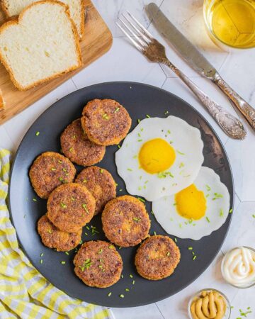 Nine round flat crispy brown sausage patties on a black plate served with fried egg look-alikes. Condiments, bread, utensils, and a glass of juice is placed next to it.