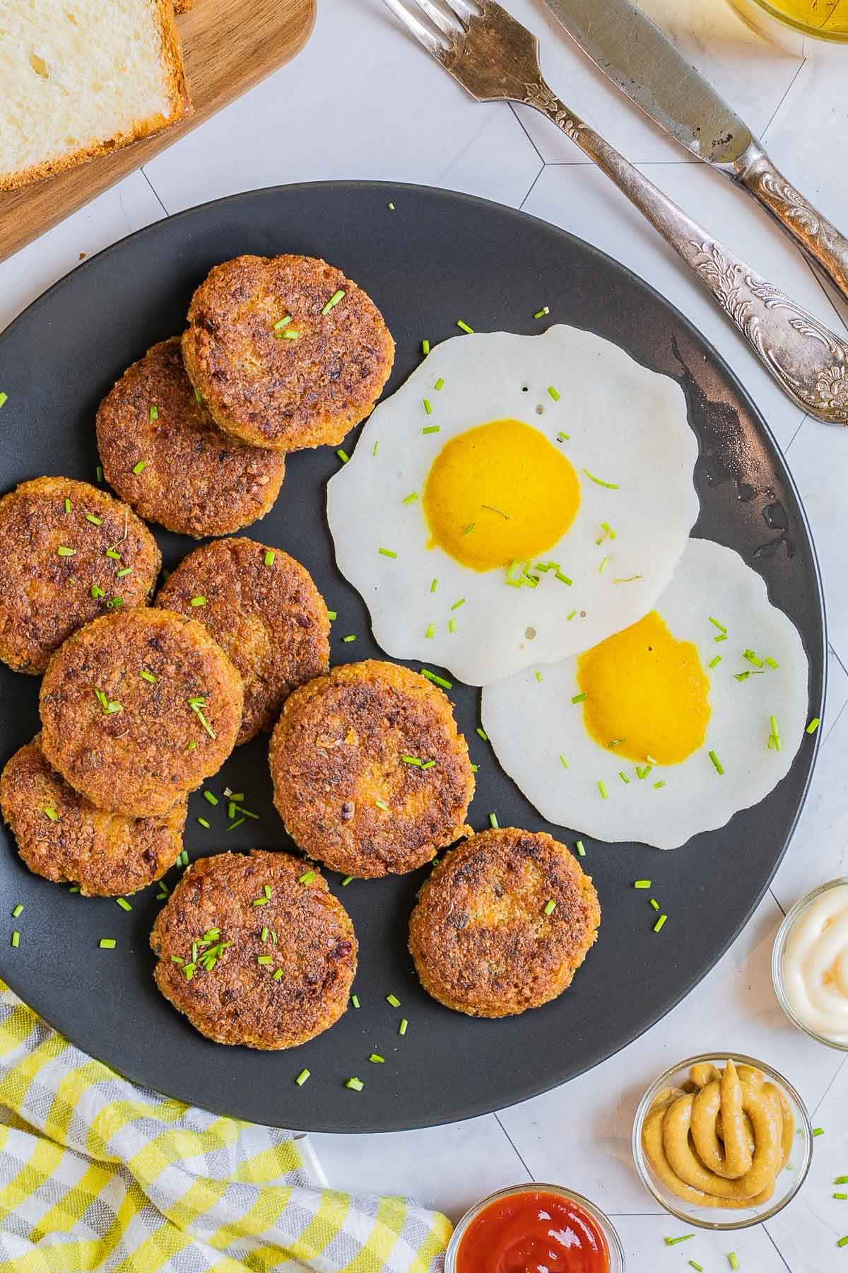 Nine round flat crispy brown sausage patties on a black plate served with fried egg look-alikes. Condiments, bread, utensils, and a glass of juice is placed next to it.