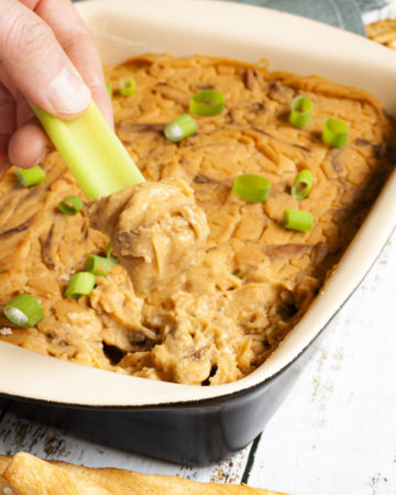 A hand is holding a green celery stick that was dipped in an orange dip with brown shreds. The dip is at the background in a light brown square dish.