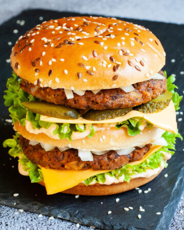 A burger with layers of green ruffled lettuce, yellow sauce, thin cheese slices, dark brown burger patty twice, sliced pickles and white chopped onion.