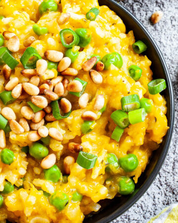 A black bowl from above on a grey surface full of yellow risotto topped with pine nuts and chopped spring onion.