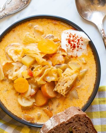 Black bowl with orange soup, sausage, carrot slices, diced potatoes and chopped cabbage topped with white sour cream. A slice of bread is placed next to the bowl as well as two spoons.