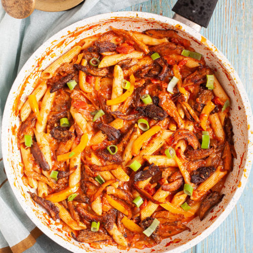 Penne pasta with yellow bell pepper slices, brown mushroom shreds and spring onion rings in a creamy red sauce in a frying pan from above.
