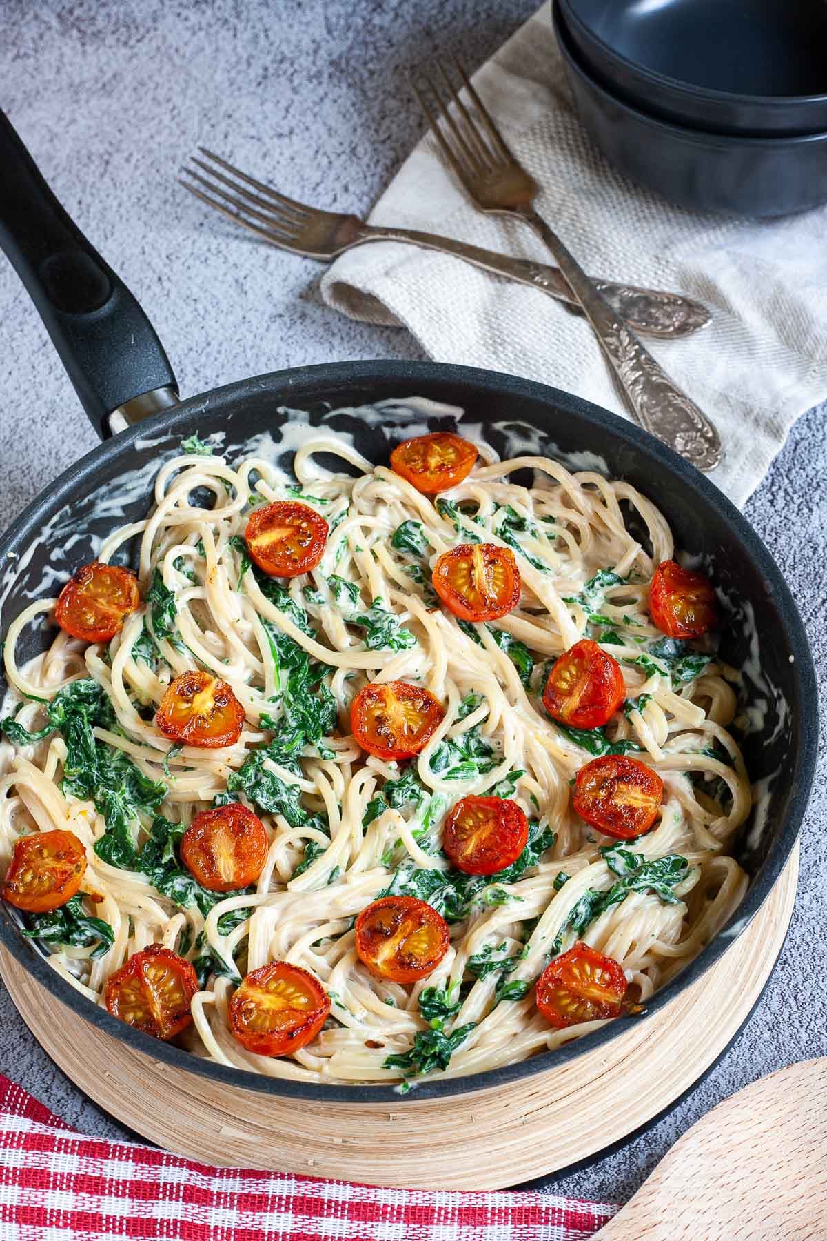 Black frying pan with spaghetti in white sauce with wilted spinach leaves and charred cherry tomatoes.