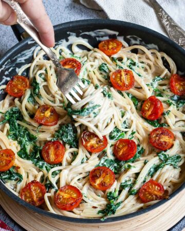 Black frying pan with spaghetti in white sauce with wilted spinach leaves and charred cherry tomatoes. A hand is holding a fork and coil up with pasta.