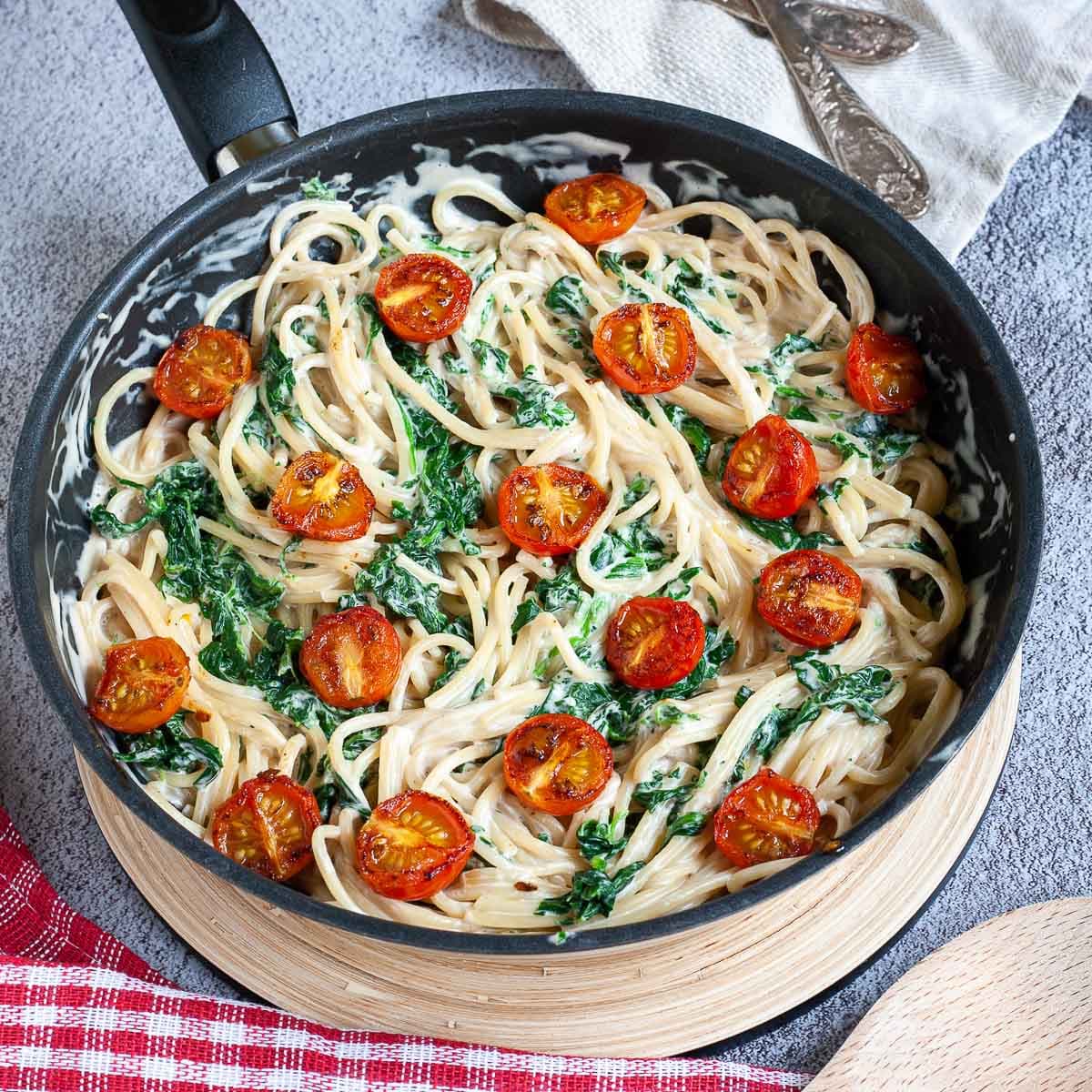 Black frying pan with spaghetti in white sauce with wilted spinach leaves and charred cherry tomatoes.