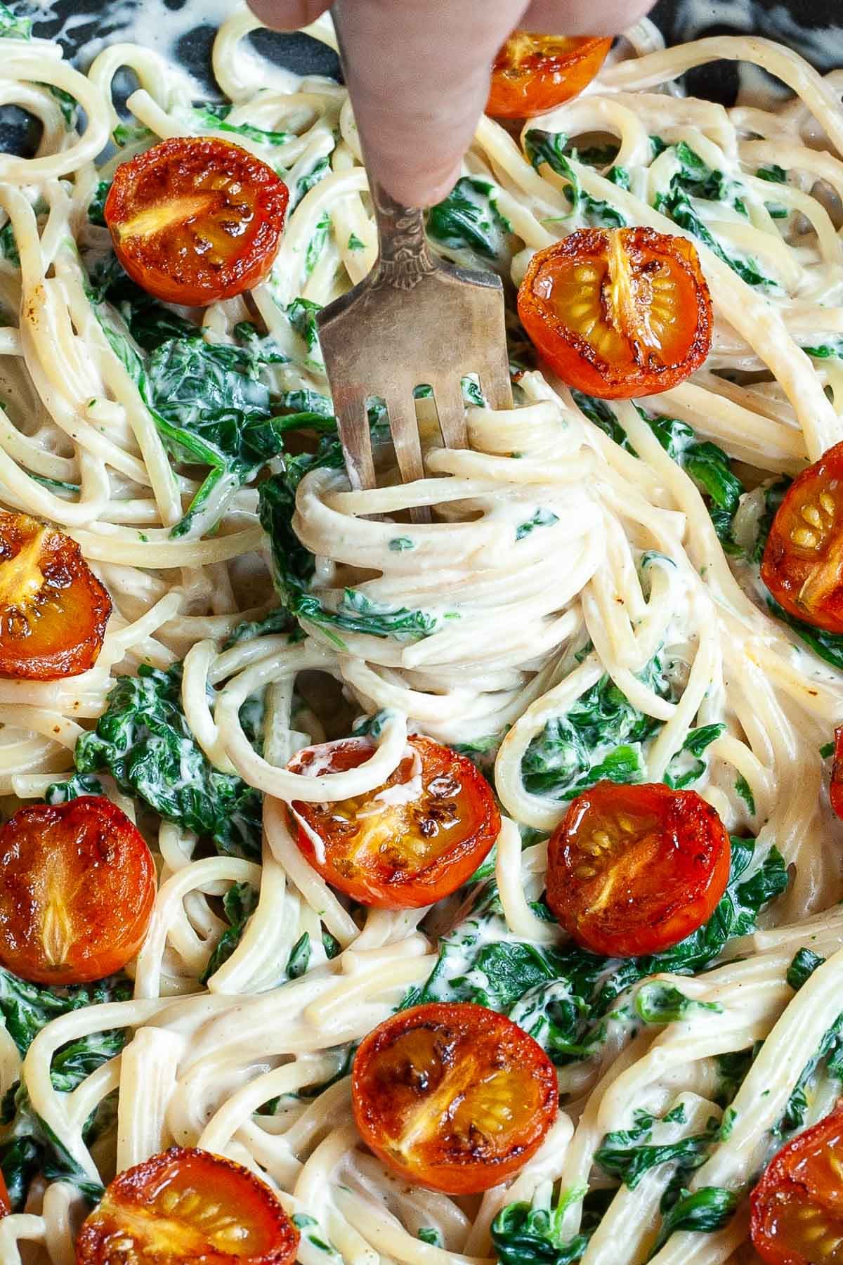 Black frying pan with spaghetti in white sauce with wilted spinach leaves and charred cherry tomatoes. A hand is holding a fork and coil up with pasta.