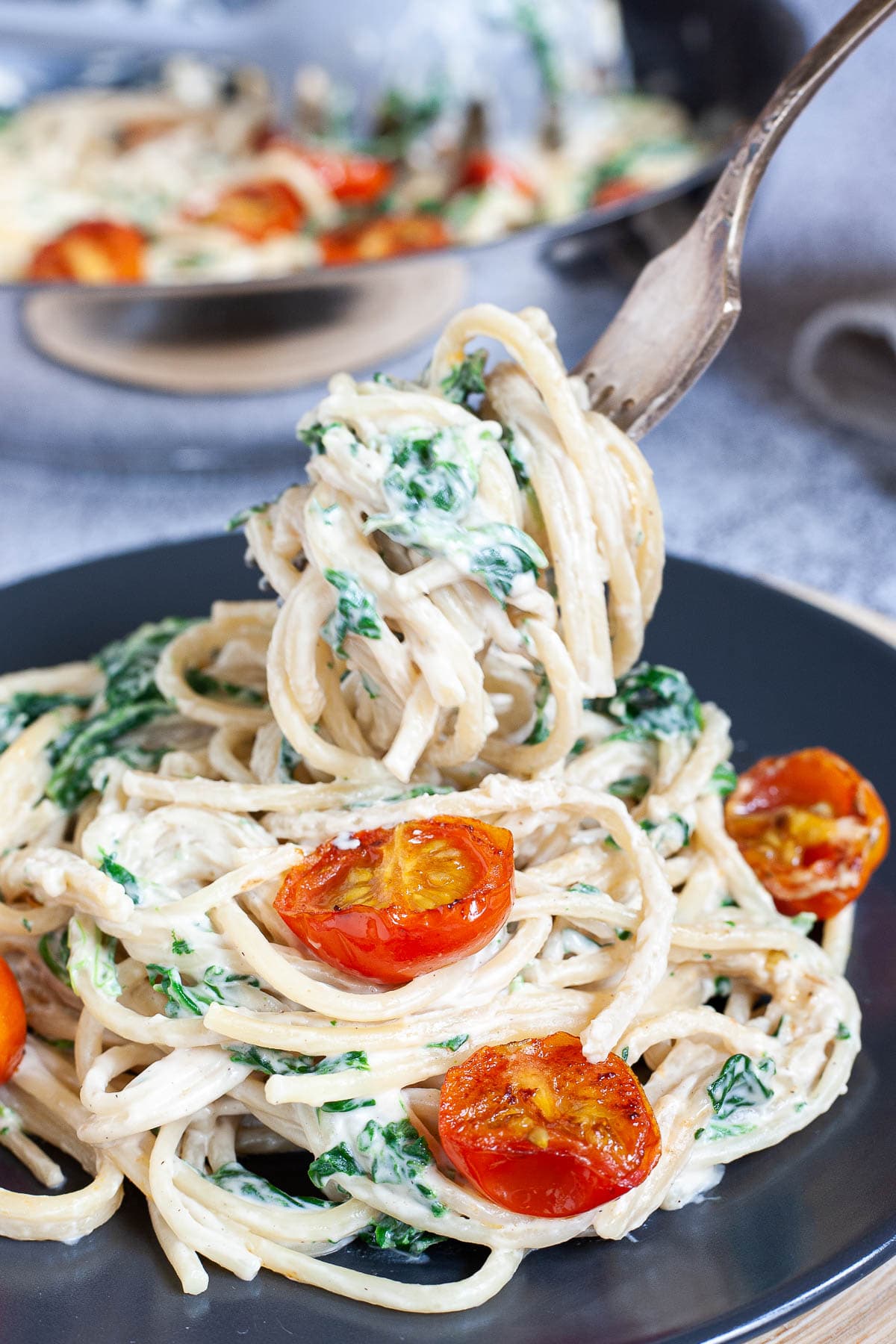 Spaghetti in white sauce is served on a black plate with charred cherry tomatoes and wilted spinach leaves. A fork is lifting some pasta up.