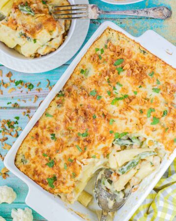 A baking dish with penne pasta in creamy sauce, chickpeas and green peas topped with breadcrumbs and freshly chopped herbs. The corner part is missing and served in a small plate on the side.