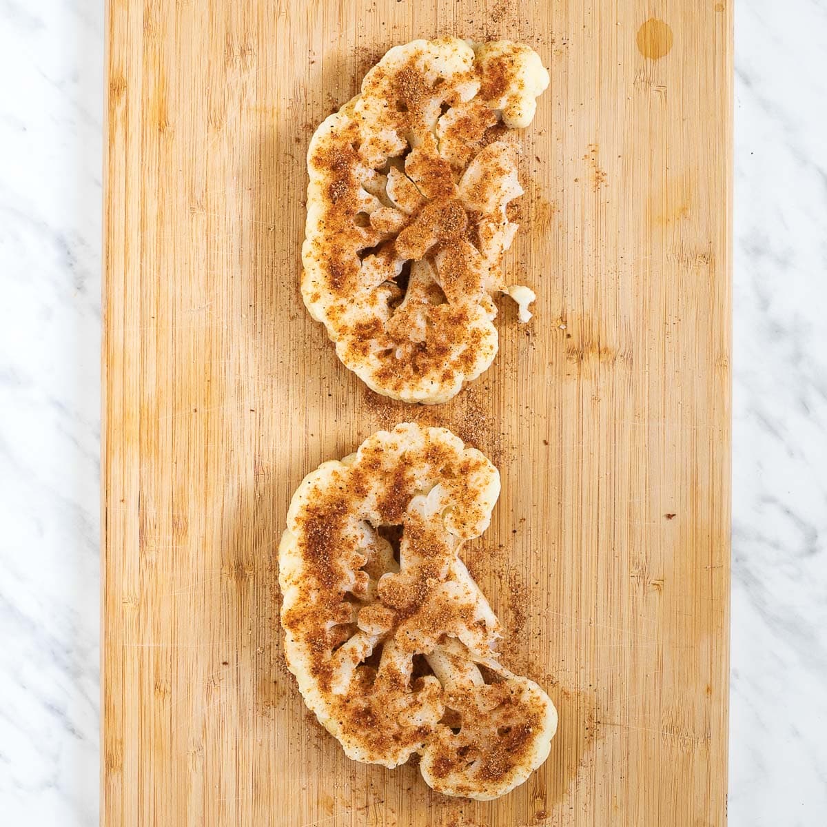 2 large cauliflower slices with ample dry spice rub on a wooden cutting board.