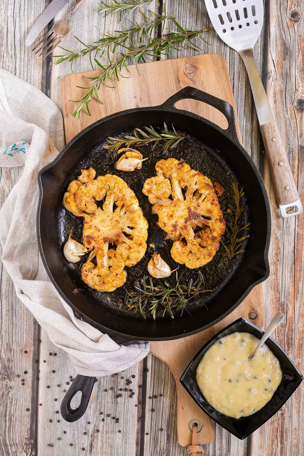 2 crispy brown yellow cauliflower slices in a cast-iron skillet surrounded by roasted garlic cloves and wilted rosemary twigs. A small black bowl of green peppercorn sauce is next to it.