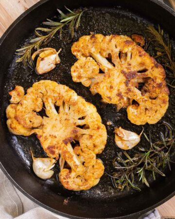 2 crispy brown yellow cauliflower slices in a cast-iron skillet surrounded by roasted garlic cloves and wilted rosemary twigs.