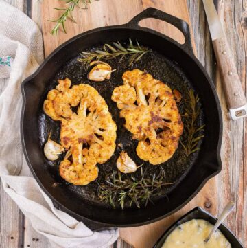 2 crispy brown yellow cauliflower slices in a cast-iron skillet surrounded by roasted garlic cloves and wilted rosemary twigs. A small black bowl of green peppercorn sauce is next to it.