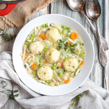 A white plate with dumplings, chopped veggies, shredded mushroom pieces in place of chicken, green herbs in a thick, white stew. The remaining soup is in a red-white enameled Dutch oven next to it.