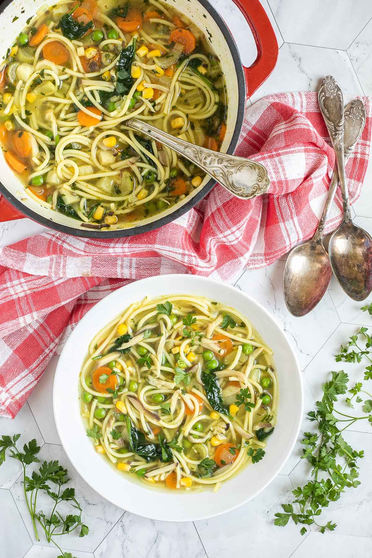 A white plate with noodles, spinach leaves, green peas, carrots slices in a thin soup. Leftover with red Dutch oven is next to it.