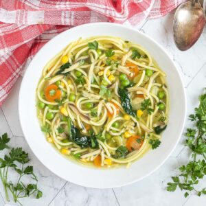 A white plate with noodles, spinach leaves, green peas, carrots slices in a thin soup.