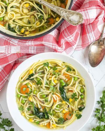 A white plate with noodles, spinach leaves, green peas, carrots slices in a thin soup. Leftover with red Dutch oven is next to it.