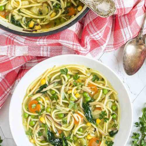 A white plate with noodles, spinach leaves, green peas, carrots slices in a thin soup. Leftover with red Dutch oven is next to it.