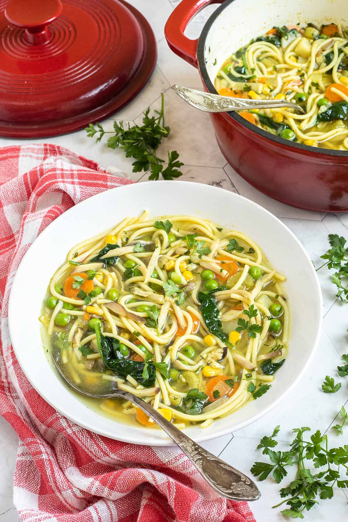 A white plate with noodles, spinach leaves, green peas, carrots slices in a thin soup. Leftover with red Dutch oven is next to it.