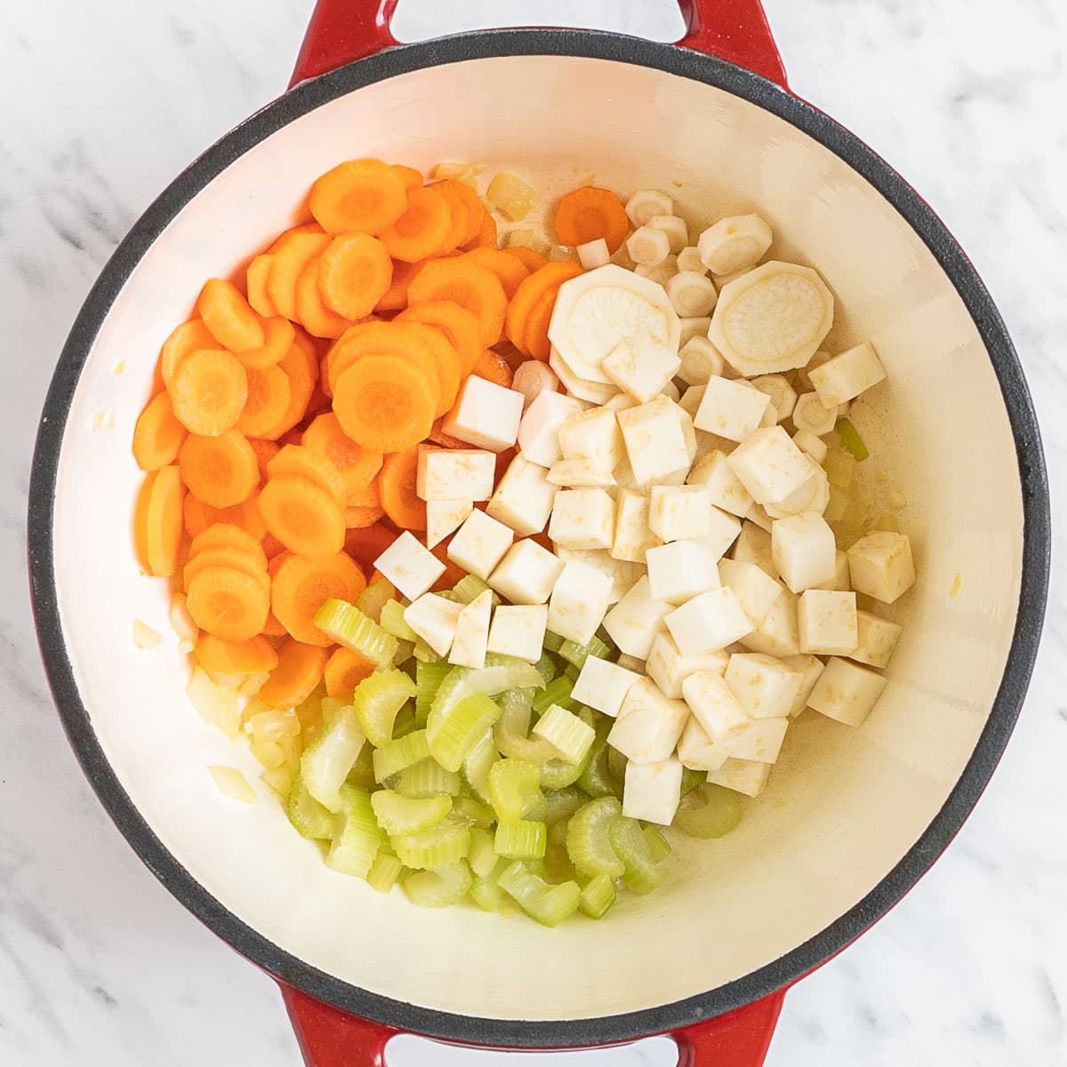 Red Dutch oven with sliced carrots, chopped parsnip, chopped celeriac, and celery stalk.