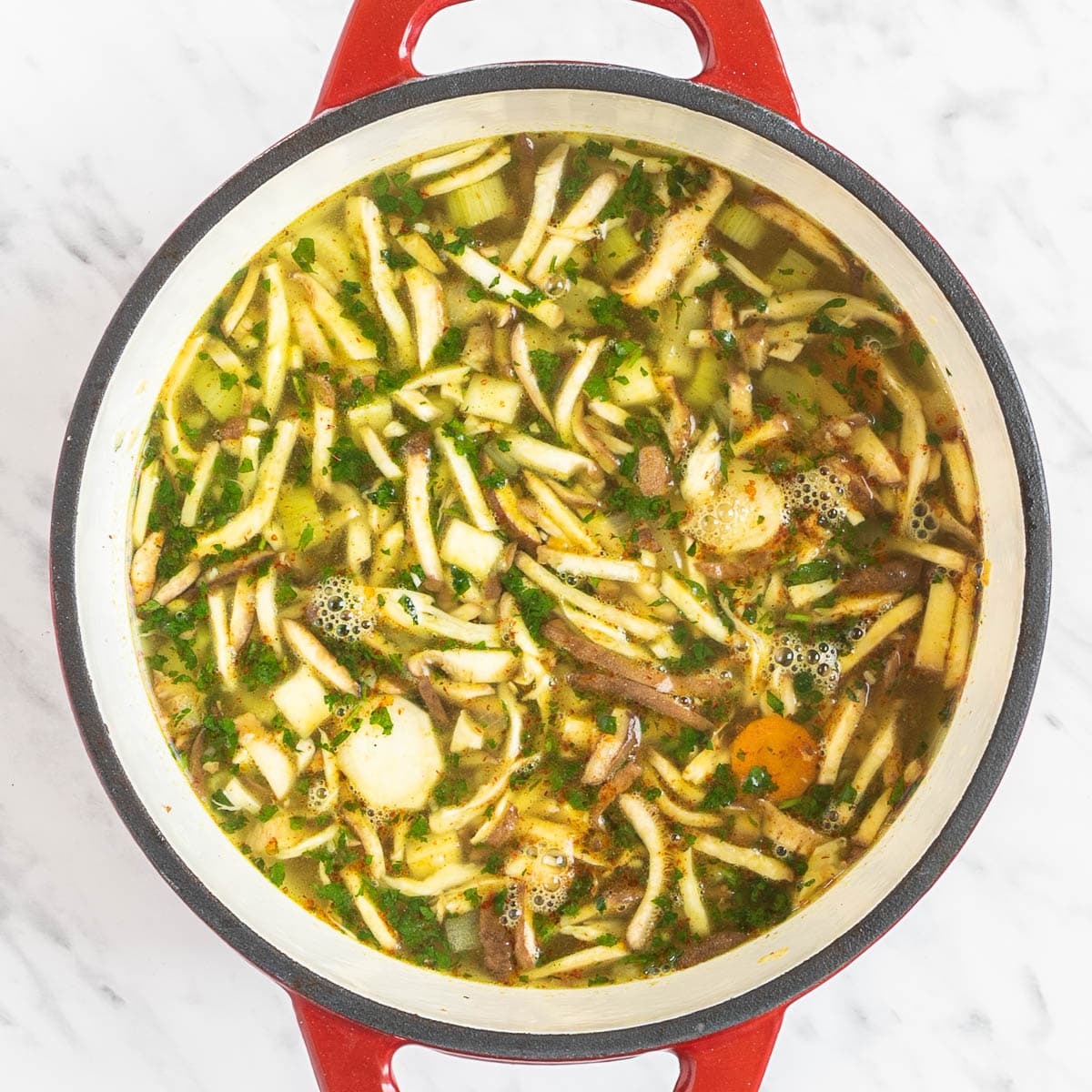 Red Dutch oven with sliced carrots, chopped parsnip, chopped celeriac, celery stalk, shredded oyster mushrooms, chopped herbs in a thin vegetable broth-like soup.