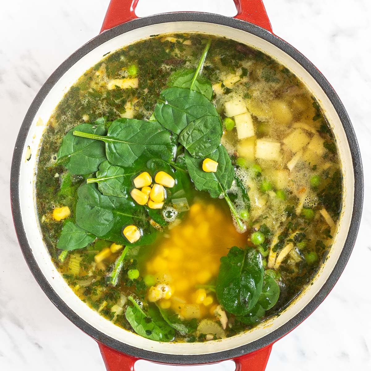Red Dutch oven with sliced carrots, chopped parsnip, chopped celeriac, celery stalk, shredded oyster mushrooms, chopped herbs, fresh spinach leaves, corn, green peas in a thin vegetable broth-like soup.