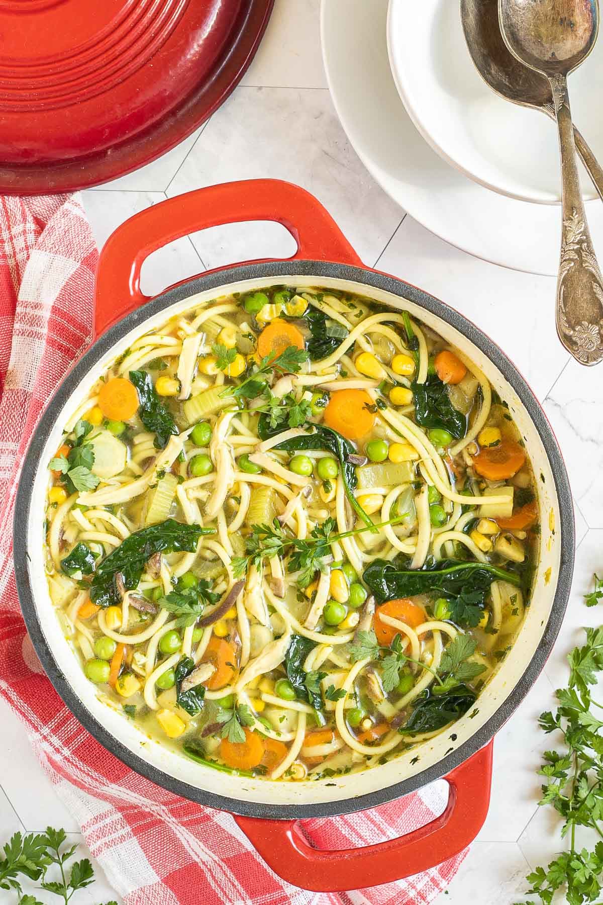Red Dutch oven with noodles, spinach leaves, green peas, carrots slices in a thin soup.