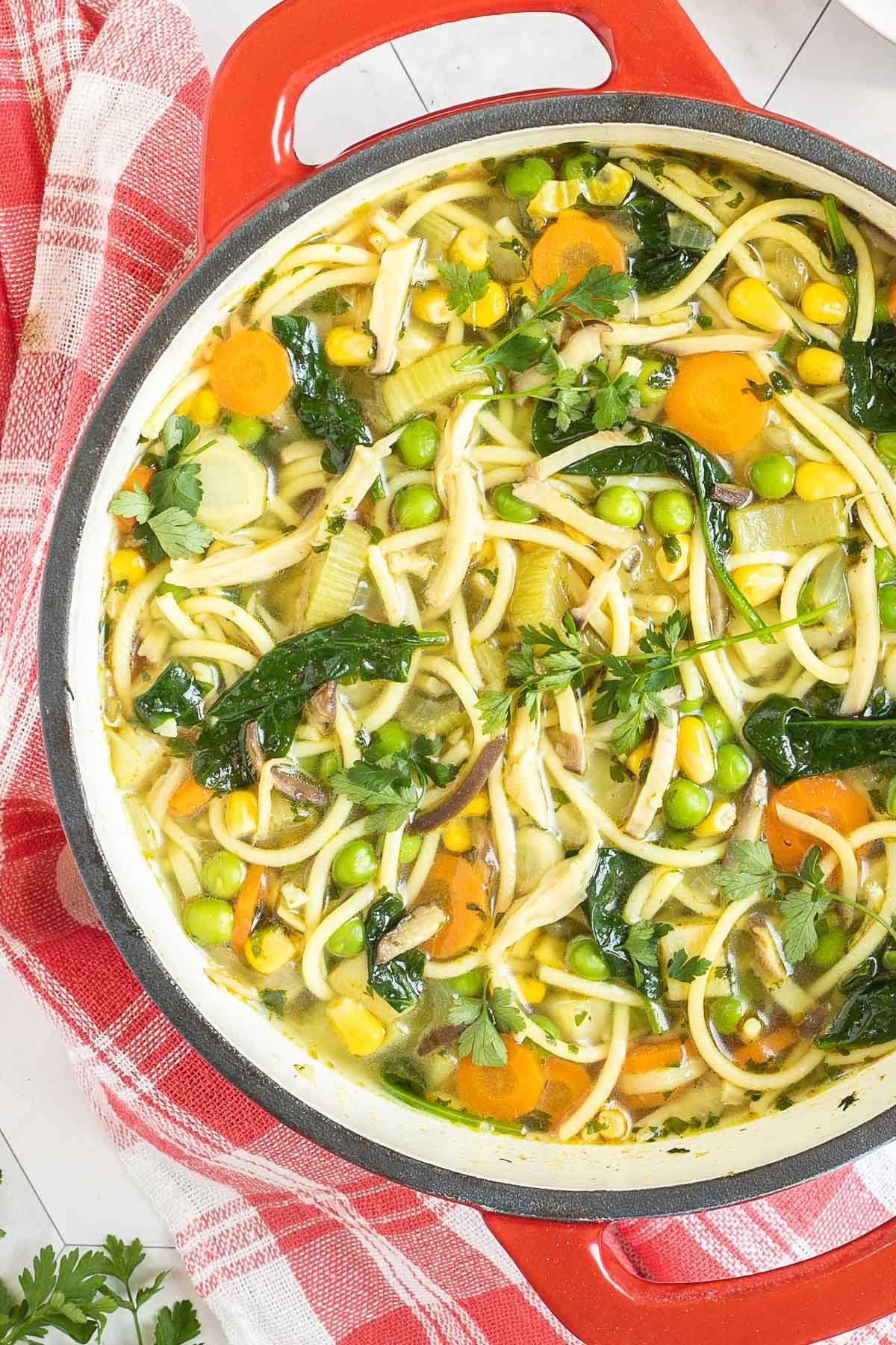 Red Dutch oven with noodles, spinach leaves, green peas, carrots slices in a thin soup.