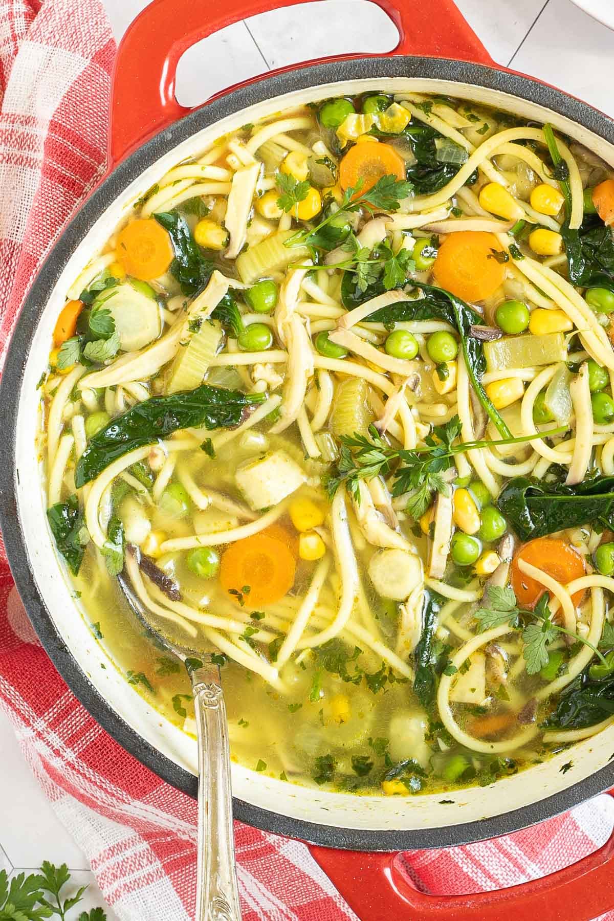 Red Dutch oven with noodles, spinach leaves, green peas, carrots slices in a thin soup.