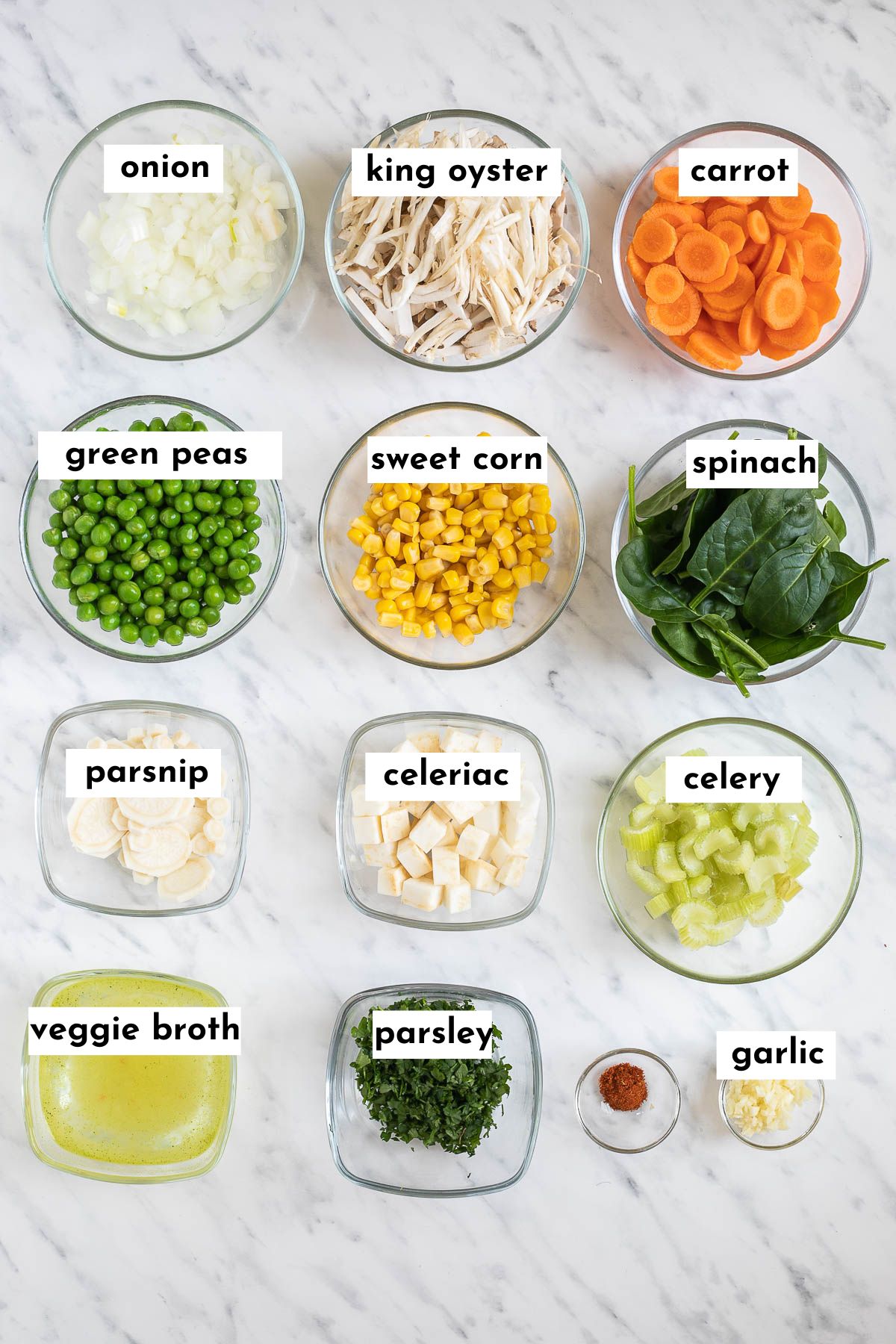 A glass bowl with the ingredients of the vegan chicken noodle soup like chopped onion, shredded mushrooms, sliced carrots, green peas, corn, spinach leaves, parsnip, celeriac, celery, veggie broth, parsley.