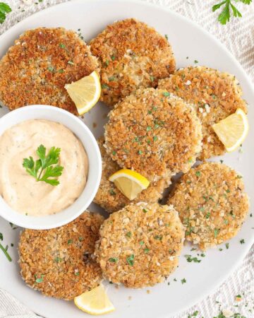 Several round-shaped breadcrumbs covered patties on a large white plate with a light orange sauce in a white bowl. Chopped parsley and lemon wedges are around for decoration.