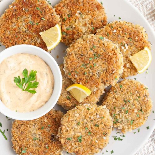Several round-shaped breadcrumbs covered patties on a large white plate with a light orange sauce in a white bowl. Chopped parsley and lemon wedges are around for decoration.