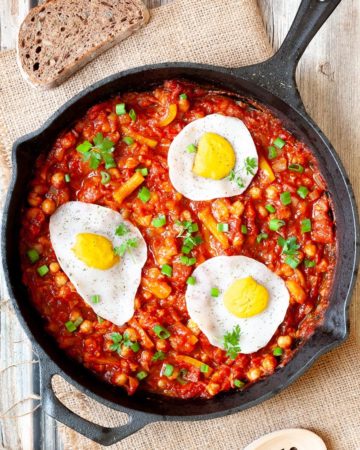 A cast iron skillet from above. 3 fried egg lookalikes with white and yolk part on top of a red chunky sauce sprinkled with green spring onion rings and chopped parsley. You can see chickpeas, chopped tomato and chopped yellow bell peppers.