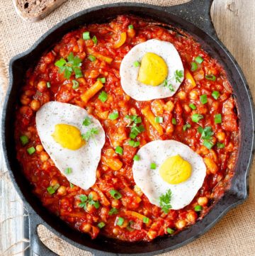 A cast iron skillet from above. 3 fried egg lookalikes with white and yolk part on top of a red chunky sauce sprinkled with green spring onion rings and chopped parsley. You can see chickpeas, chopped tomato and chopped yellow bell peppers.