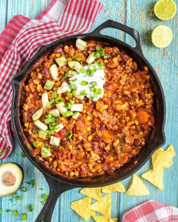 Cast iron skillet with a chunky chili loaded with chopped vegetables and beans, topped with white cream, chopped avocado, and scallion. Half avocados and tortilla chips are around it.