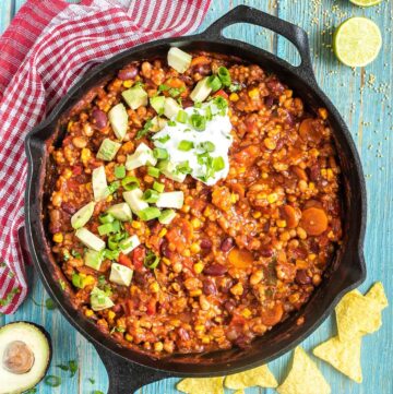 Cast iron skillet with a chunky chili loaded with chopped vegetables and beans, topped with white cream, chopped avocado, and scallion.