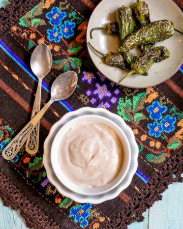 Light orange thick dip is in a small bowl with roasted green peppers on the side. Placed on a colorful brown, blue, green woven tablecloth.