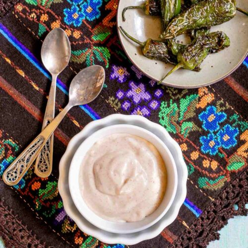Light orange thick dip is in a small bowl with roasted green peppers on the side. Placed on a colorful brown, blue, green woven tablecloth.