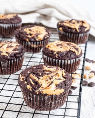 Several chocolate muffins with a peanut butter swirl on top on a black wire rack. Even more chocolate chips and peanuts are scattered around and under the rack.