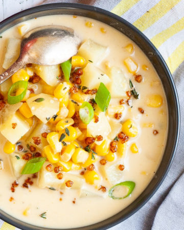 Light yellow soup served in a black bowl with croutons, corn, and diced potatoes. It is sprinkled with freshly chopped herbs, brown bacon bits and sliced spring onion.