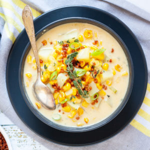 Light yellow soup served in a black bowl with croutons, corn, and diced potatoes. It is sprinkled with freshly chopped herbs, brown bacon bits and sliced spring onion.