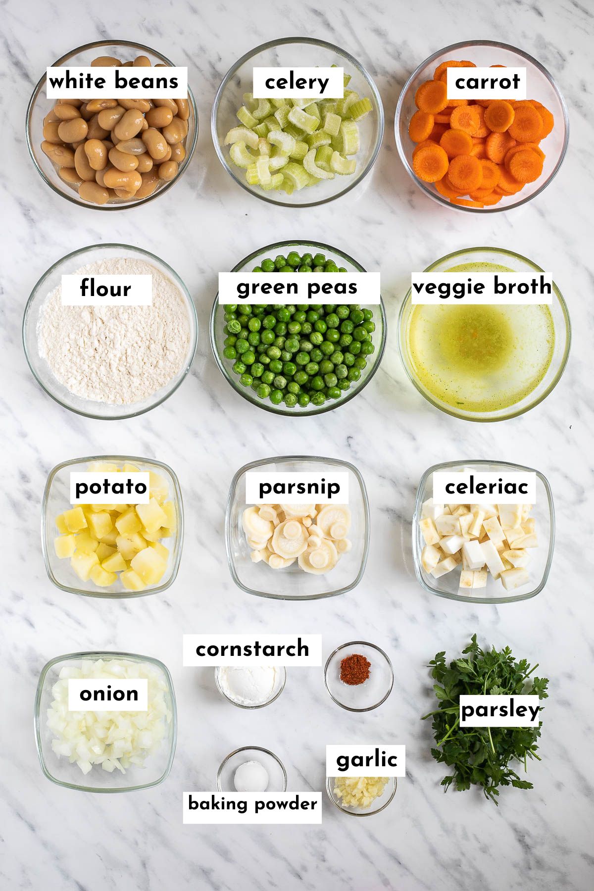 The ingredients of vegan dumpling soup are in small glass bowls like white beans, green peas, chopped celery, sliced carrots, chopped potatoes, chopped parsnip, celeriac root, flour, veggie broth, onion and fresh green herbs