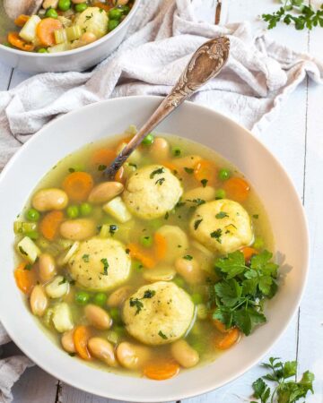 Two white plates with dumplings, chopped veggies, green herbs, white beans, and green peas in a vegetable broth soup. A spoon is placed in it.