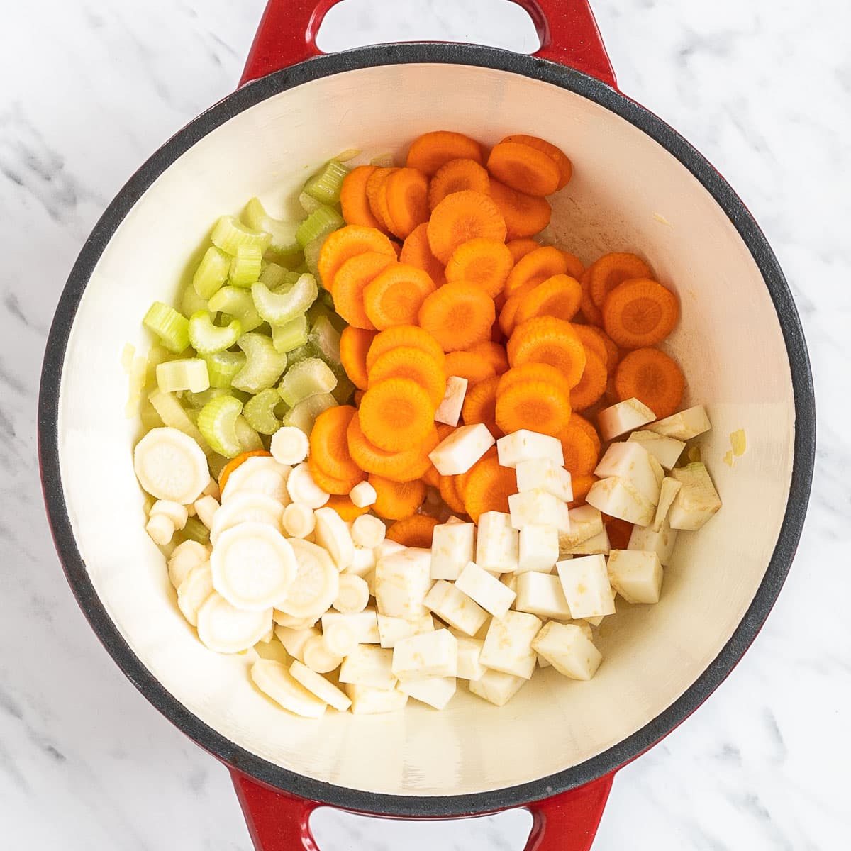 Red white enameled Dutch oven with chopped carrots, celery stalks, celeriac root and parsnip,